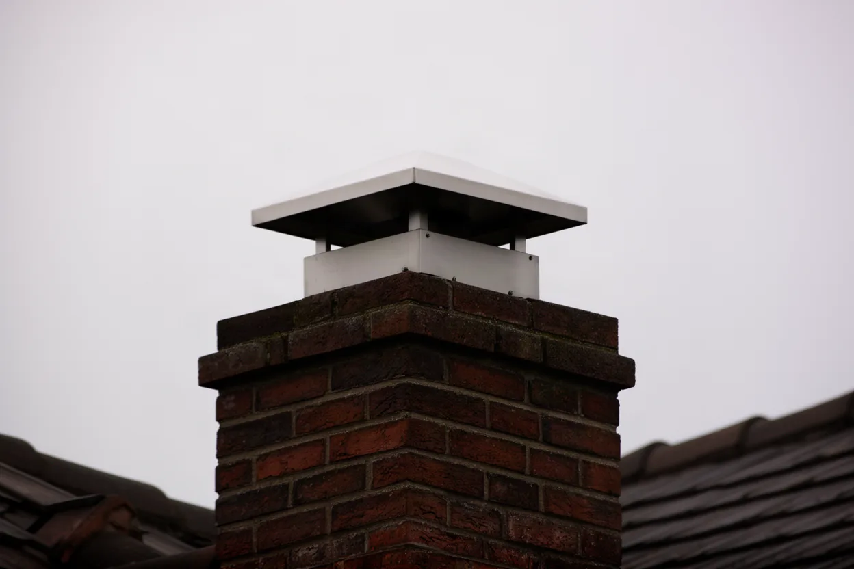 a stainless chimney cap on a brick chimney against the sky, rooftop visible, muted colors